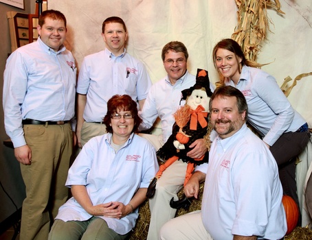 Six people in light blue shirts pose together indoors with a decorative orange and black puppet.