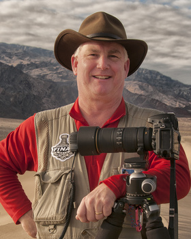 Photographer holding telephoto lens on tripod mount against mountainous landscape backdrop.