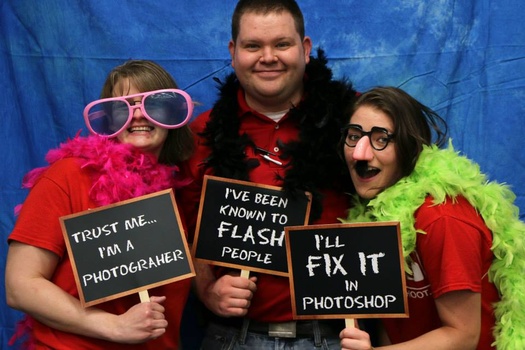 Three people in colorful wigs holding signs with photography jokes against a blue backdrop.