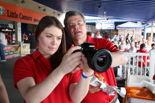 Two people in red shirts holding a DSLR camera at an indoor event venue.