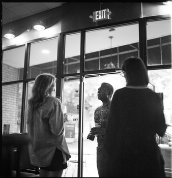 Three people viewed from behind looking out through glass doors with an illuminated exit sign above.