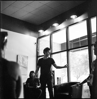 Man standing by large windows in an interior space with overhead lighting, seated figure visible in foreground.