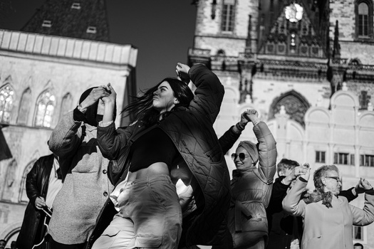 A group of people looking upward in an urban square, some shielding their eyes, with historic architecture in the background.