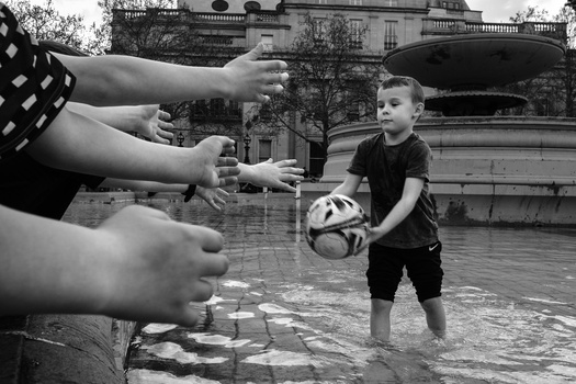 Child holding a soccer ball in shallow water while adults' hands reach toward him from the left edge.