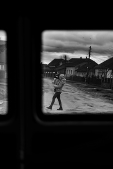 Black and white photograph framed through a window, showing a person walking on a wet street with buildings and cloudy sky.