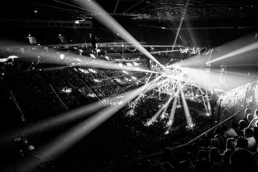Dramatic overhead view of a concert venue with powerful spotlight beams cutting through darkness and thousands of audience members.