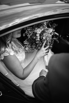 Bride holding bouquet in car, groom's hand visible beside her in black and white photograph.