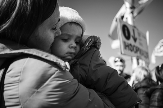 Woman holding a young child at what appears to be an outdoor gathering or protest, both warmly dressed.