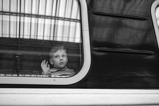 Young child looking out through a rounded window of a vehicle, hand pressed against the glass.