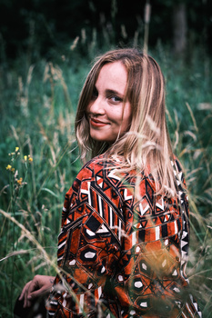 Young woman with long blonde hair wearing a patterned red jacket, smiling at camera in a grassy field.