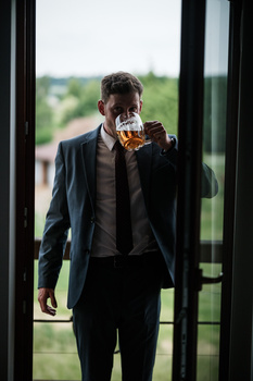 Man in dark suit and tie holding whiskey glass while standing by large windows overlooking green landscape.