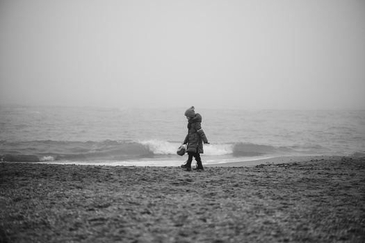Child in winter coat standing alone on pebbly beach facing calm, misty ocean.