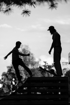 Two silhouetted figures in business attire stand on a wooden structure against a bright sky with tree branches overhead.