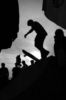 Skateboarder mid-air performing a trick against a dramatic cloudy sky, silhouetted with spectators below.