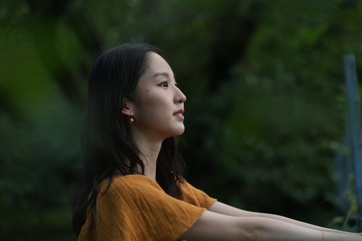 Woman in golden-yellow shirt gazing upward against blurred green foliage background.