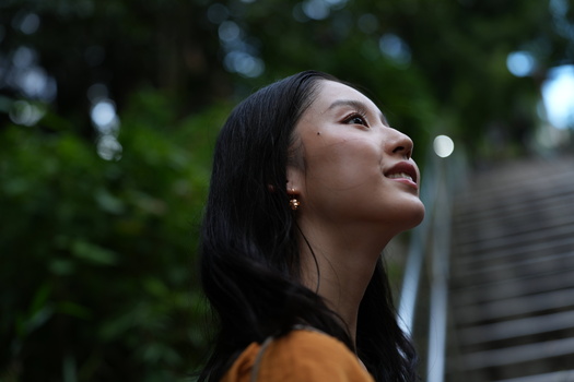 Woman with long dark hair looking upward, wearing an orange-yellow top against a blurred green outdoor background.