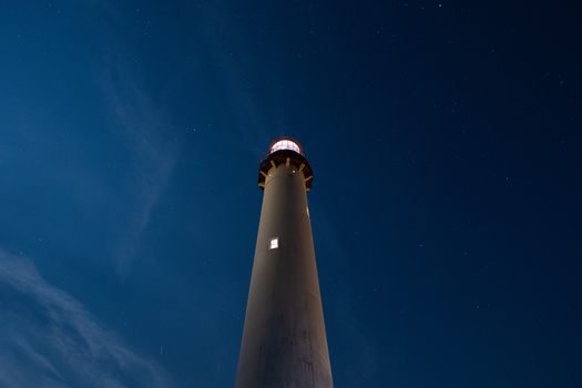 Tall lighthouse tower shot from below against a deep blue sky with wispy clouds and visible stars.