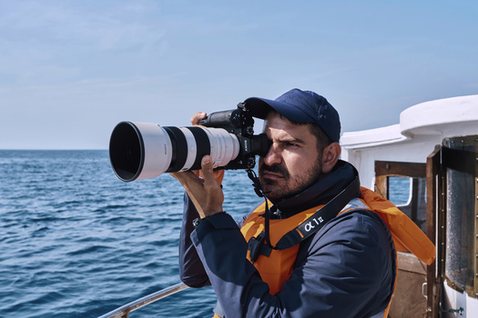 Photographer holding telephoto lens while shooting from a boat on calm ocean water.