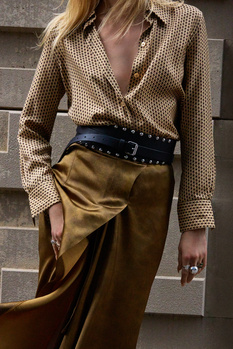 Woman wearing a cream polka dot blouse and olive silk skirt with a studded black belt.