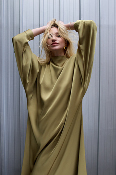 Woman in oversized olive green sweater with hands raised to head against corrugated metal wall.