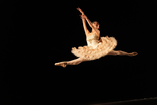 Ballet dancer performing a grand jeté in a cream-colored tutu against a black background.