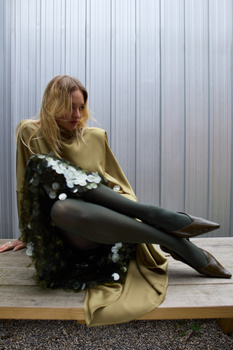 Woman in mustard coat seated against corrugated metal backdrop, holding black feathered garment.