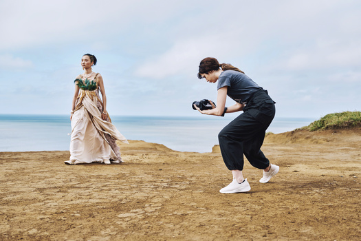 Photographer crouching with camera while model in beaded dress poses on beach during daytime.