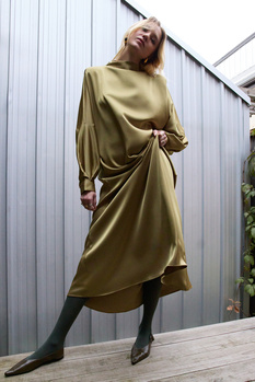 Woman in olive dress and black tights standing against corrugated metal wall.