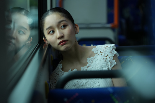 Young woman in white lace clothing looking toward camera with contemplative expression, with blurred reflections visible in glass.