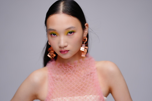 Woman with bold yellow eyeshadow and pink lipstick wearing textured pink sleeveless top against neutral background.