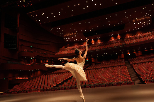 Ballet dancer in white tutu performing grand jeté on stage in empty arena with red seating.