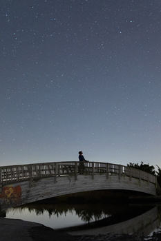 Silhouetted figure standing on arched wooden bridge at dusk with starry sky and water reflection below.
