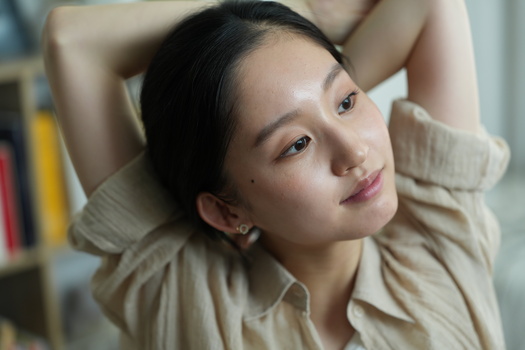 Woman in beige shirt with hands behind head, relaxed pose against blurred indoor background.