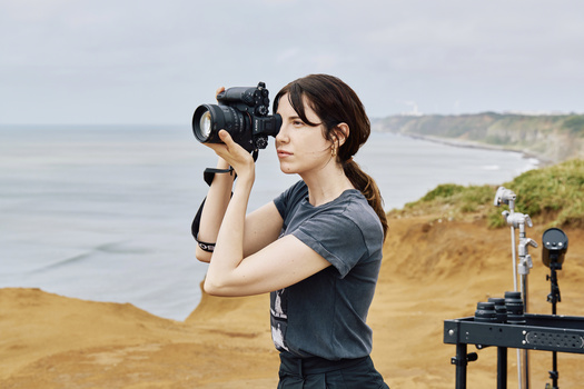 Female videographer holding a cinema camera at a coastal cliff location with ocean and landscape visible behind her.