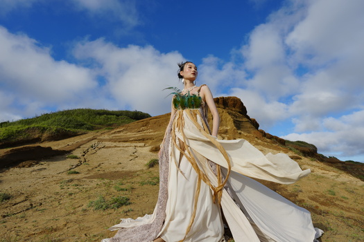 Woman in flowing white gown with green leaf crown on hillside landscape during golden hour.