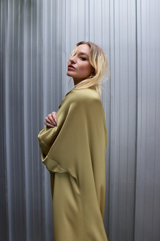 Woman in olive-toned oversized coat standing against corrugated metal wall, looking over shoulder.