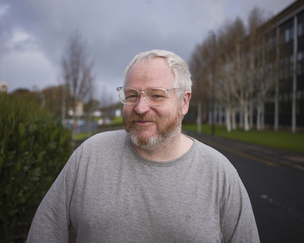Older man wearing glasses and gray shirt standing outdoors with hedges and buildings visible behind him.