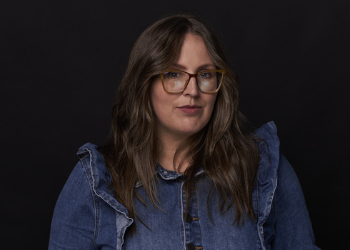 Woman wearing glasses and denim jacket against dark background.