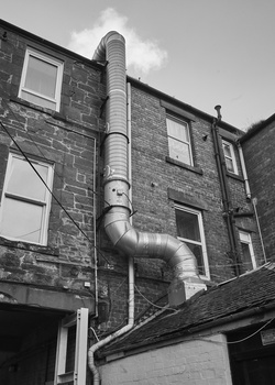 Black and white photograph of a stone building with a large metal drainage pipe running diagonally across its facade.