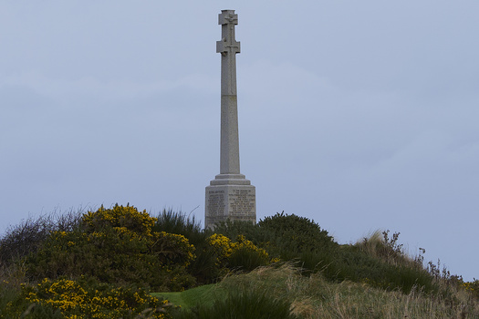 Tall stone Celtic cross monument on a grassy hilltop surrounded by yellow gorse and coastal vegetation.