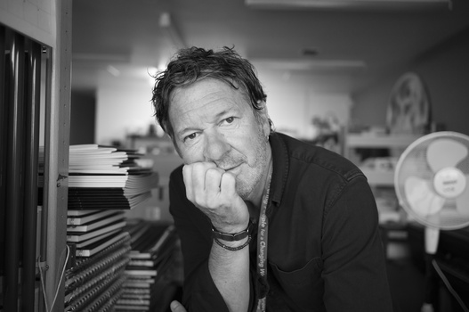 Man in dark shirt resting chin on hand, surrounded by stacked CDs and studio equipment in black and white.