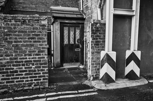 Black and white photograph of a narrow alley with brick walls, a dark doorway, and white chevron-striped bollards.