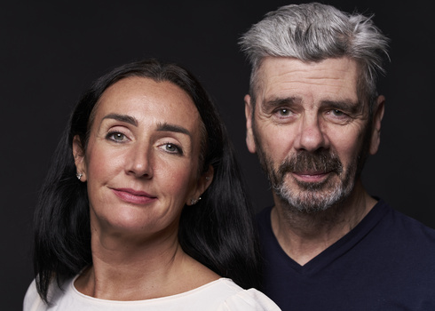 Portrait of two people against a dark background with neutral expressions.