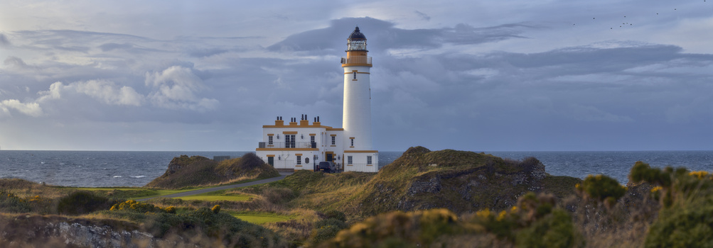 White lighthouse with golden roof section on grassy rocky peninsula surrounded by sea under overcast sky.