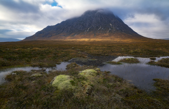 Cone-shaped mountain rising from a moorland valley with scattered pools of water and moss-covered ground beneath cloudy sky.