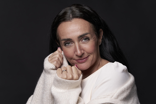 Woman with long dark hair wearing a cream-colored knit sweater against a dark background.
