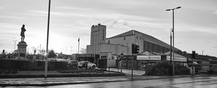 Monochrome street scene with statue, industrial buildings, and wet pavement reflecting overcast sky.