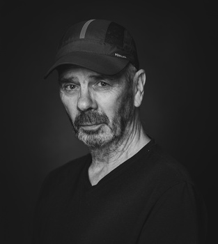 Black and white portrait of a man wearing a dark v-neck shirt and baseball cap against a dark background.