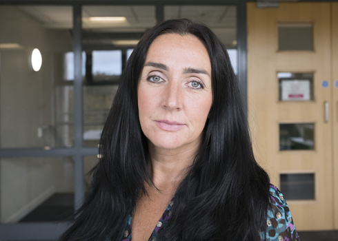 Woman with long dark hair looking directly at camera in an office hallway with glass partitions.