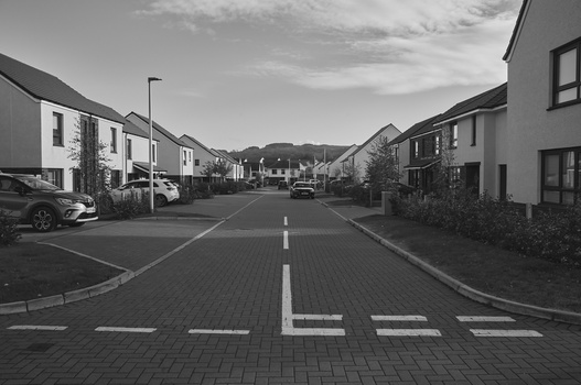Residential street with symmetrical houses, white road markings, and hills in background under cloudy sky.